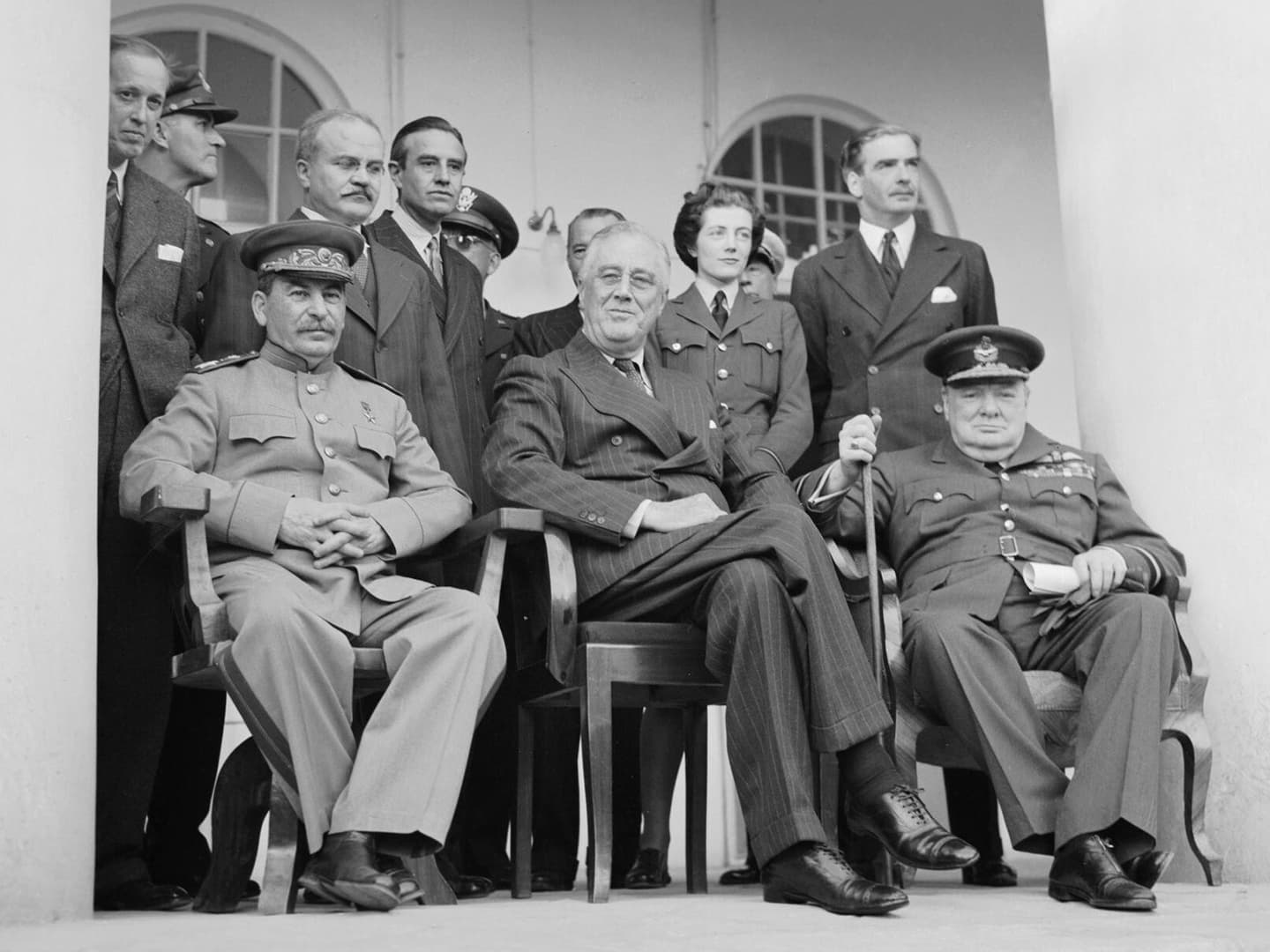 A black and white photo of a group of people between 2 white arches. Stalin, Roosevelt and Churchill sit at the front, with Spencer-Churchill standing behind.