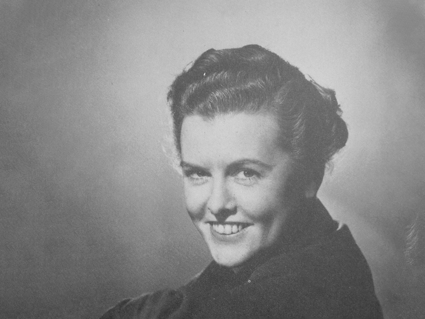 A black and white historic photo of a woman smiling at the viewer over her left shoulder.