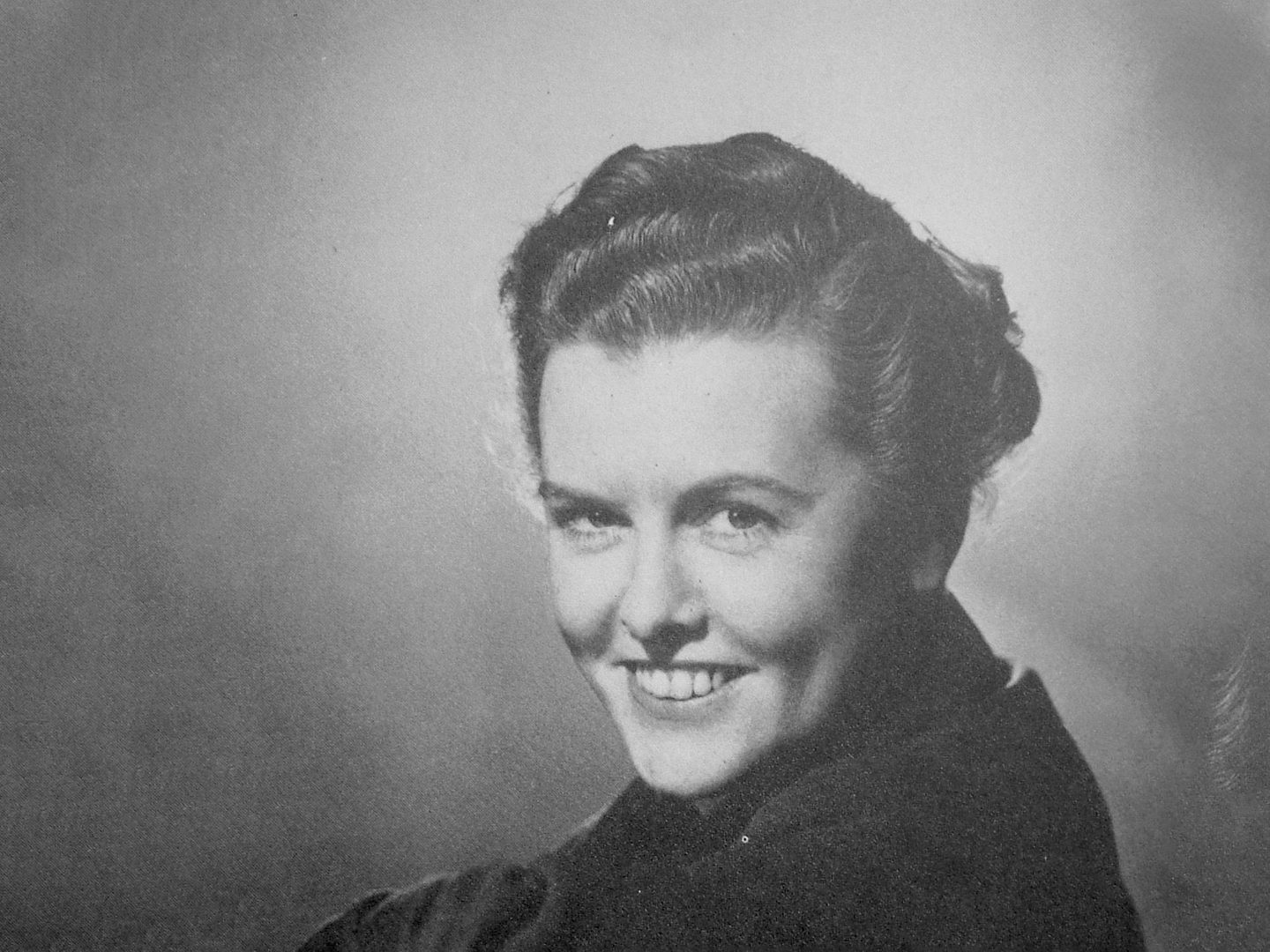 A black and white historic photo of a woman smiling at the viewer over her left shoulder.
