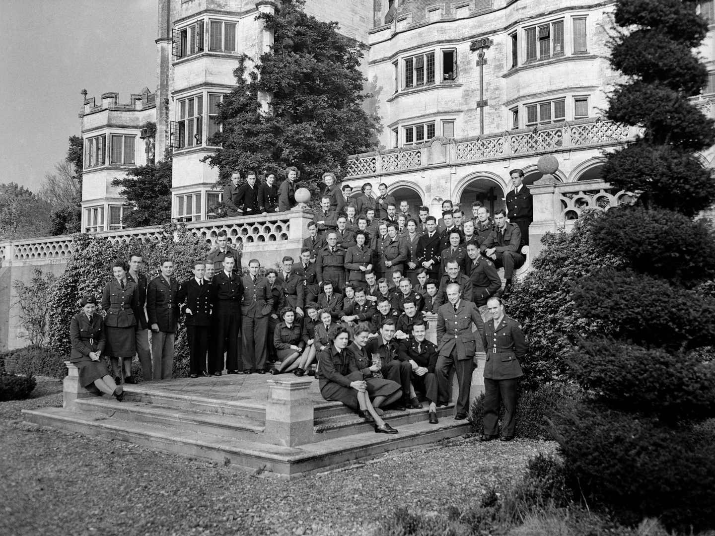 A black and white historic photo of a large group of uniformed women and men standing on some steps in front of a grand stately home. A balustrade is visible behind them, and there is a topiary bush to the right of the image.