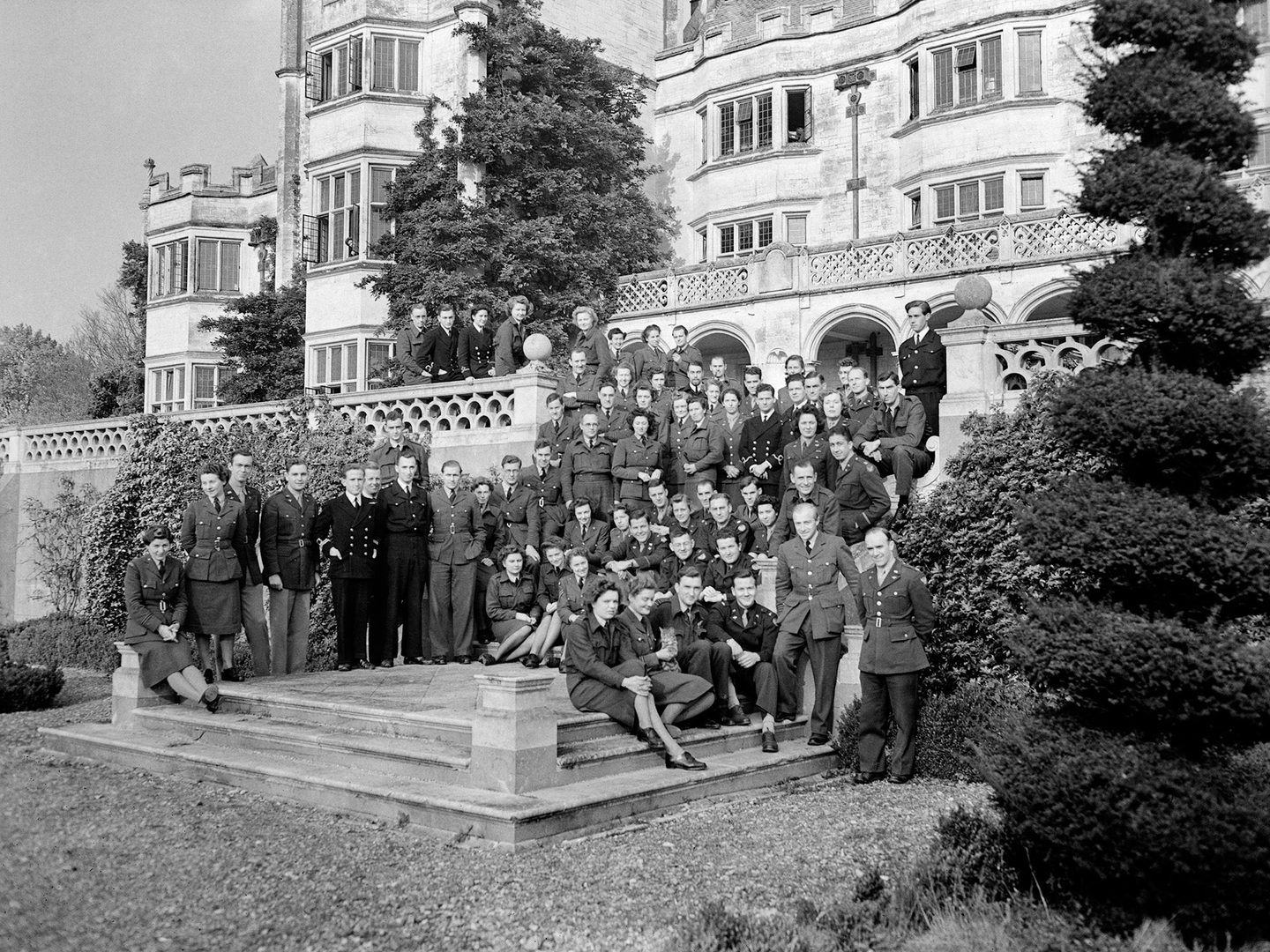A black and white historic photo of a large group of uniformed women and men standing on some steps in front of a grand stately home. A balustrade is visible behind them, and there is a topiary bush to the right of the image.