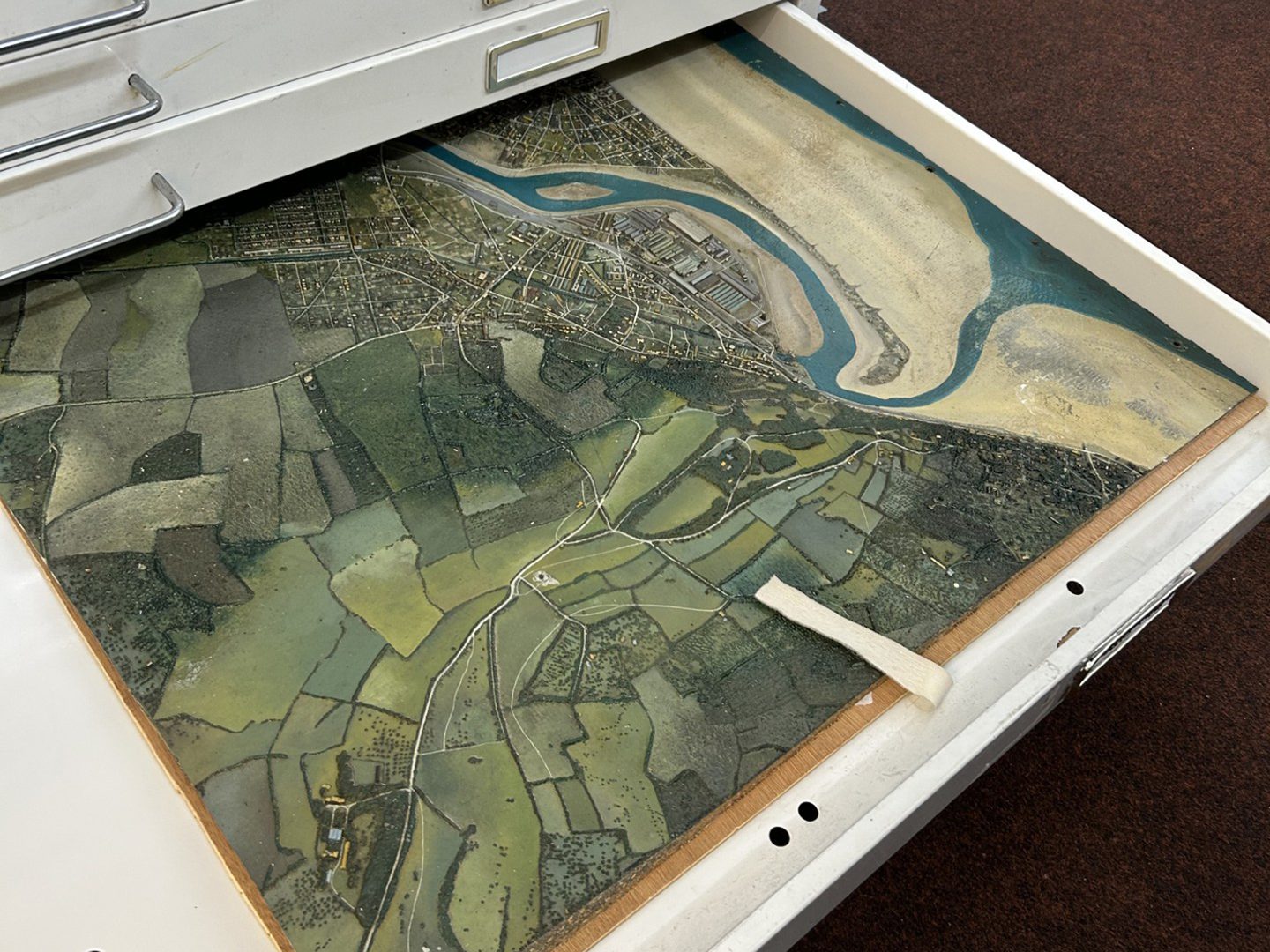 An open plan chest drawer containing a rubber model of a small town on the coast. A river runs through the town and it backed by fields. The model has been painted in naturalistic colours.