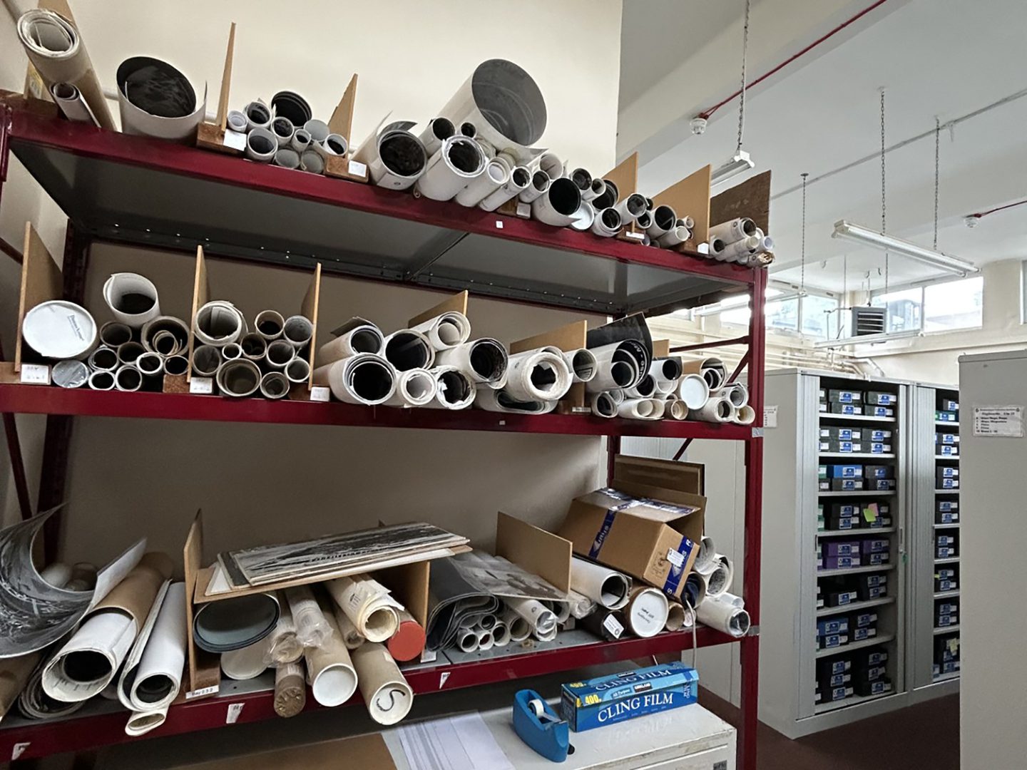 Rolls of archive material on a red shelving unit. More boxes of archival material are visible on shelves in the background.