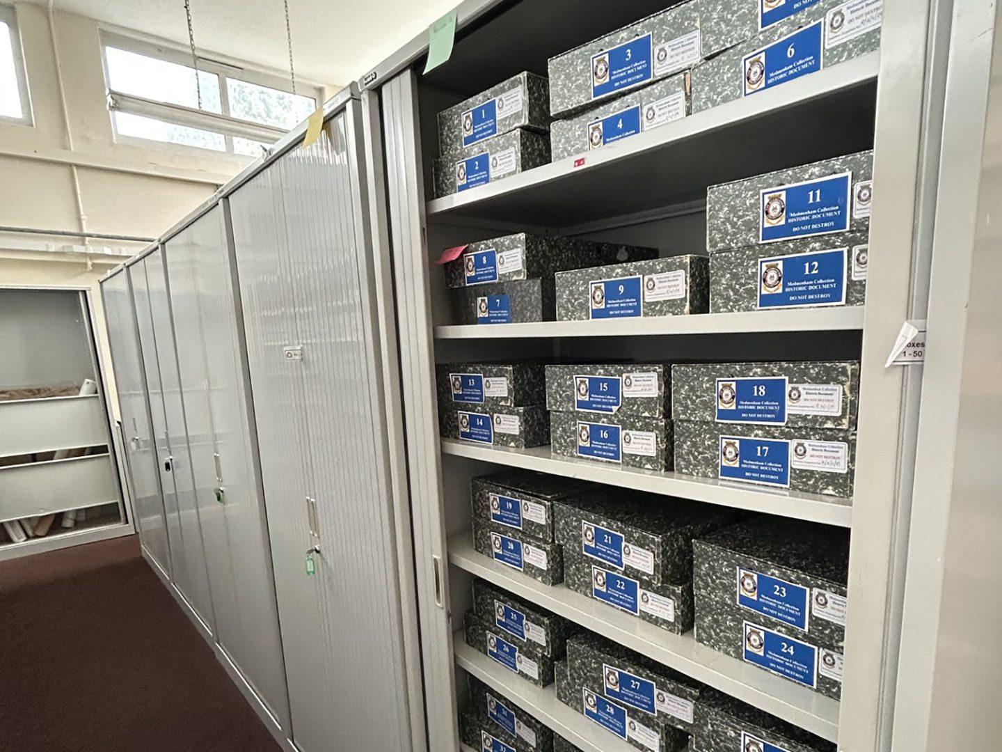 Boxes of archival material on a set of metal shelving. The shelving has doors which can be closed. More units with the doors closed are visible further along the same run of shelves.