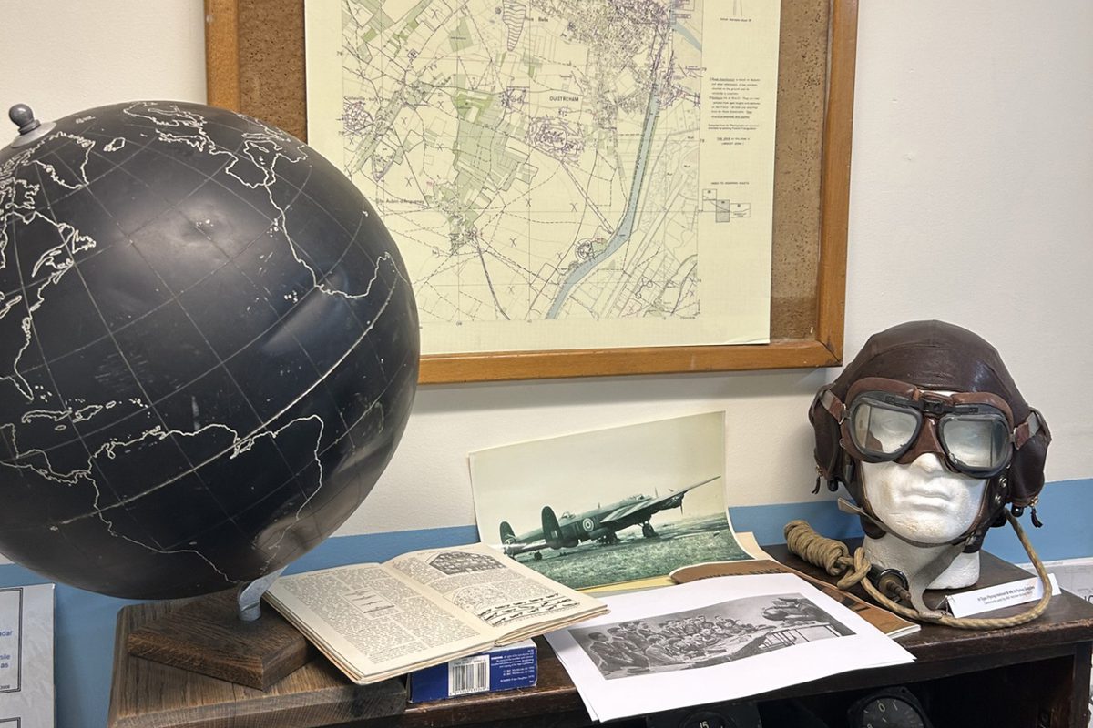 A flying cap and goggles, a photo of an aircraft, an open book and a black globe rest on a table. Behind the table is a framed map.