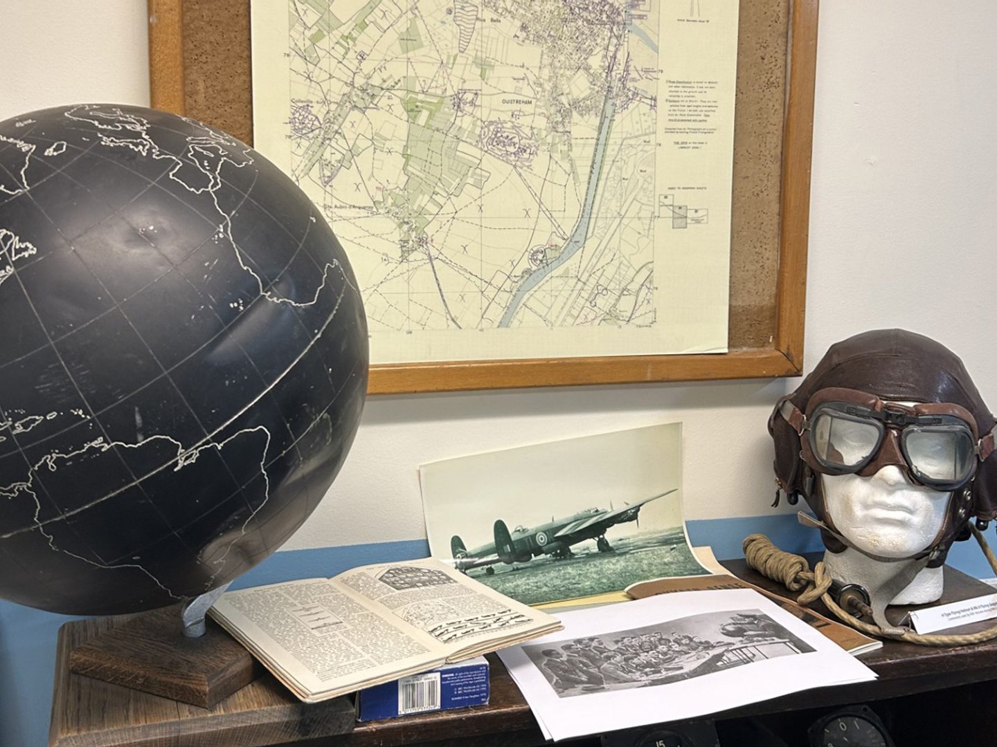 A flying cap and goggles, a photo of an aircraft, an open book and a black globe rest on a table. Behind the table is a framed map.