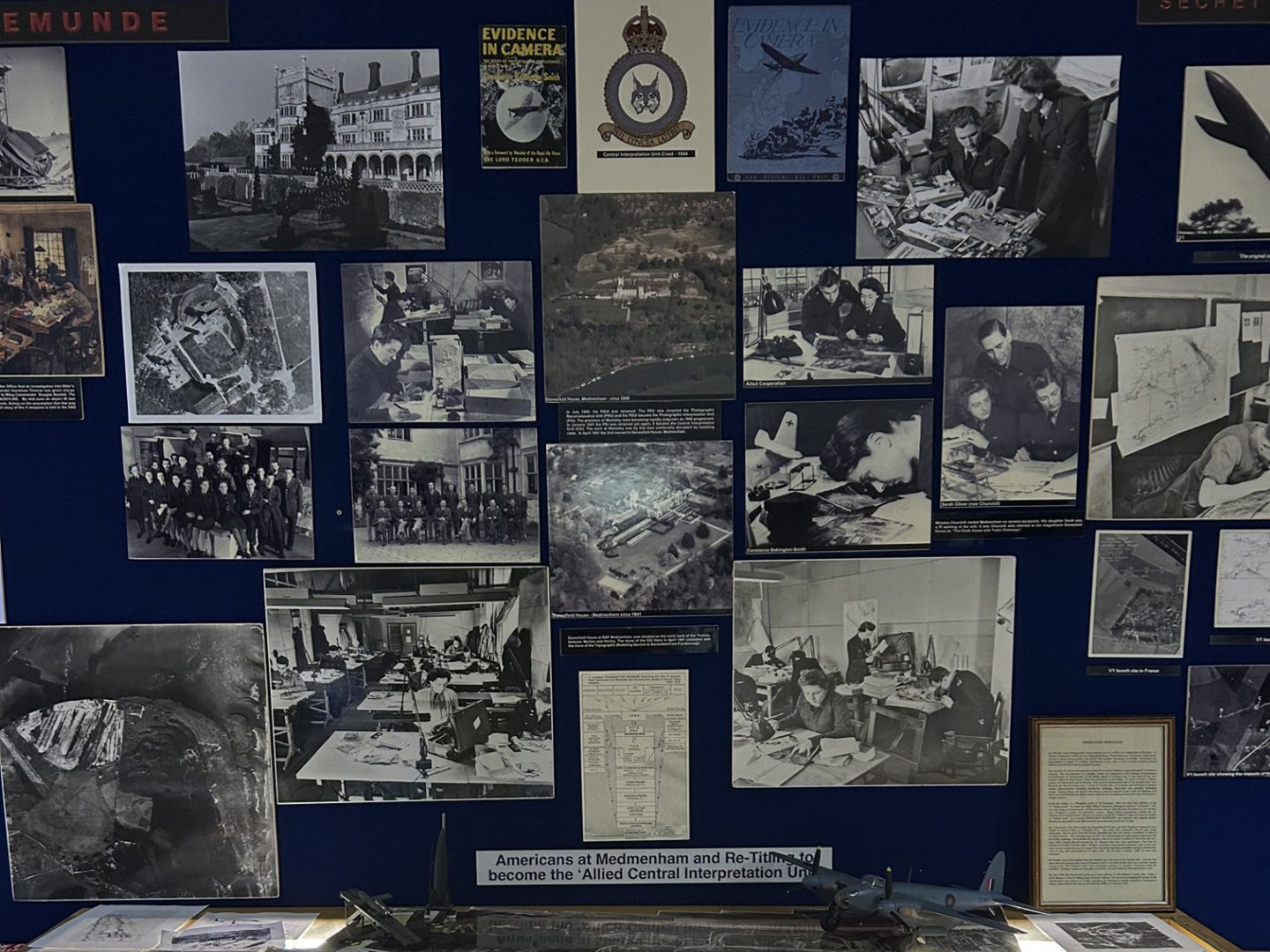 A pin board covered with photos and other imagery. The photos are mostly aerial photographs or shots of photographic interpreters at work. A model Mosquito aircraft is visible at the bottom edge.