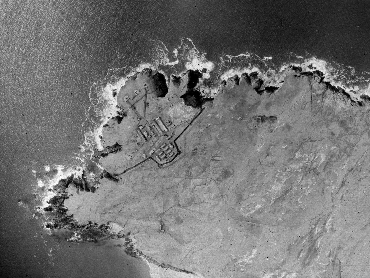 A historic black and white vertical aerial photo of a headland with cliffs surrounded by the sea on three sides. A fort is located at the tip of the headland, protected by a wall cutting it off from the mainland.