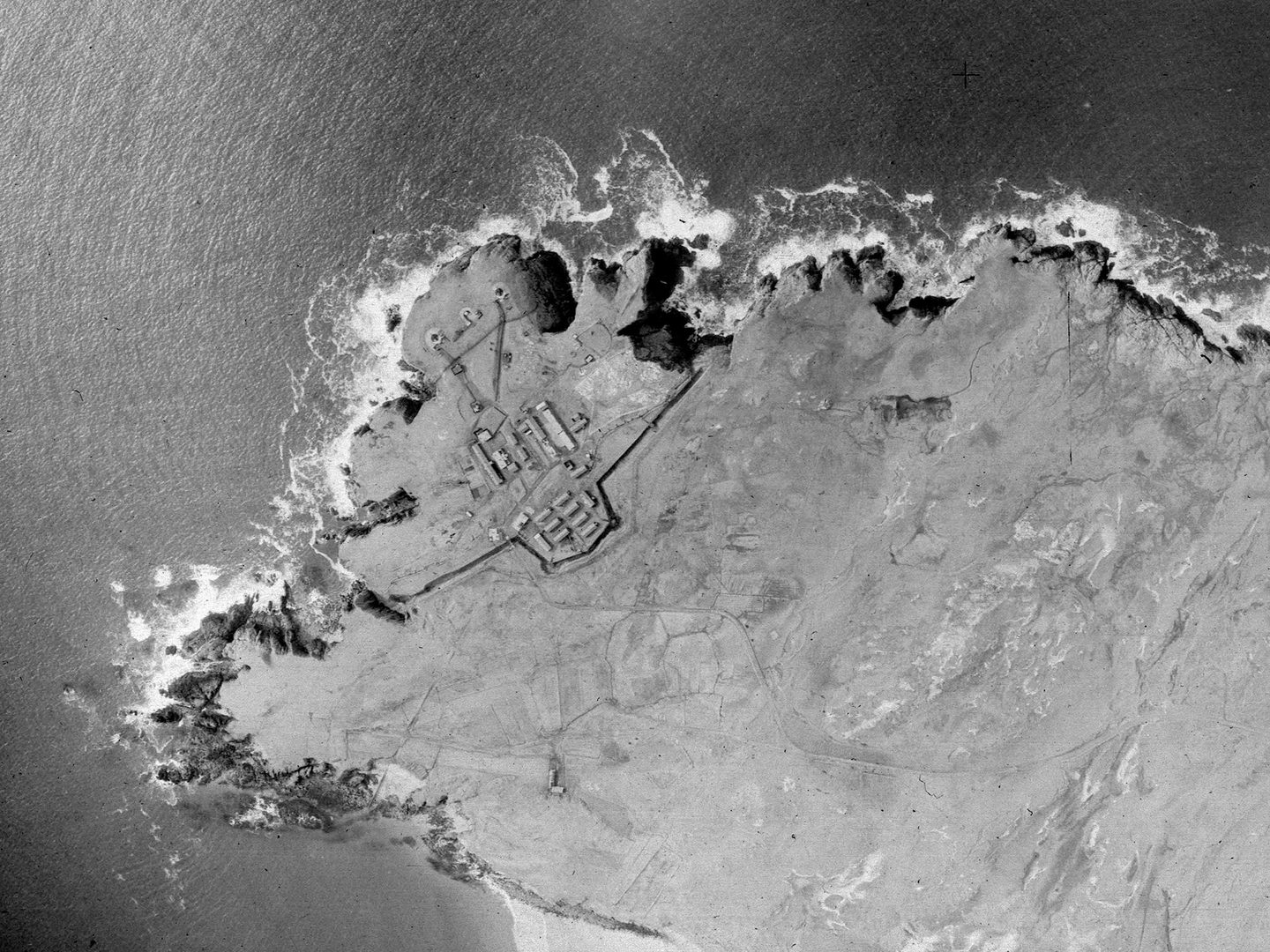A historic black and white vertical aerial photo of a headland with cliffs surrounded by the sea on three sides. A fort is located at the tip of the headland, protected by a wall cutting it off from the mainland.
