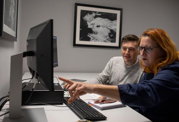 Two figures fill the right side of the frame, and sit in front of a computer workstation on the left. The figure on the right has short, dark brown hair an is wearing a grey jumper. The figure on the right has long red hair and wears glasses with a dark frame and a dark blue top. The figure on the right is pointing at the screen. Two frames black and white images are displayed on the grey walls around them.