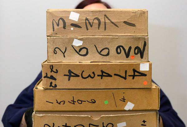 A person in a blue lab coat holds a pile of 6 small cardboard boxes. They are wearing black gloves and the top of their head is just visible above the boxes. The boxes have writing in black marker on them.