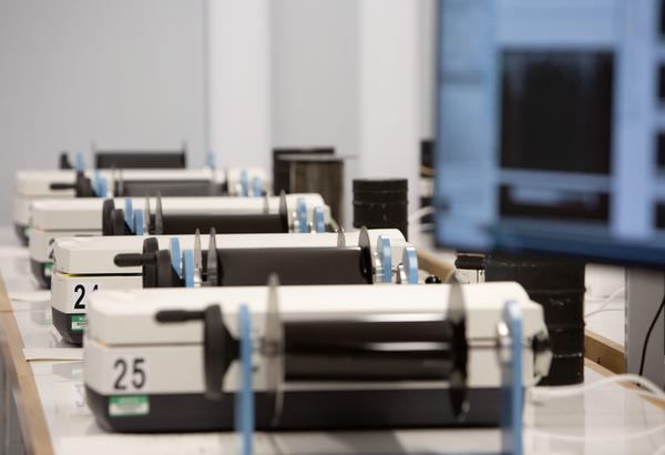 Four white and grey image scanners are spaced across a line of desks. Five blue and black film winders are spaced between the scanners. Each winder has a roll of black film on it. A section of a screen is in partial focus in the top right corner of the frame.