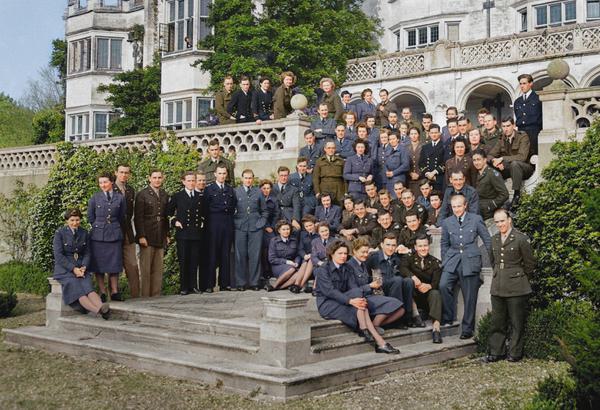 Approximately 60 figures stand in a loose group in front of a large, grey stone building. Shrubs grow on either side of the group, and appear to be climbing up the building. The figures are arranged across a stepped patio area and tiered garden. Some figures smile gently. They wear a mix of military type uniforms - there are four or five variations. For example - a navy blue fitted jacket, pencil skirt and grey tights, or a dark khaki green jacket with gold coloured buttons, and straight legged trousers.
