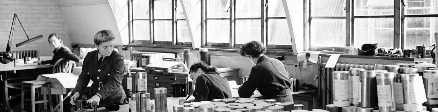 A black and white historic photo of 4 women in uniform working in an office. Large windows are visible in the background. 2 of the women face the camera, 2 are sitting with their backs to the camera. Metal film canisters cover almost every surface.