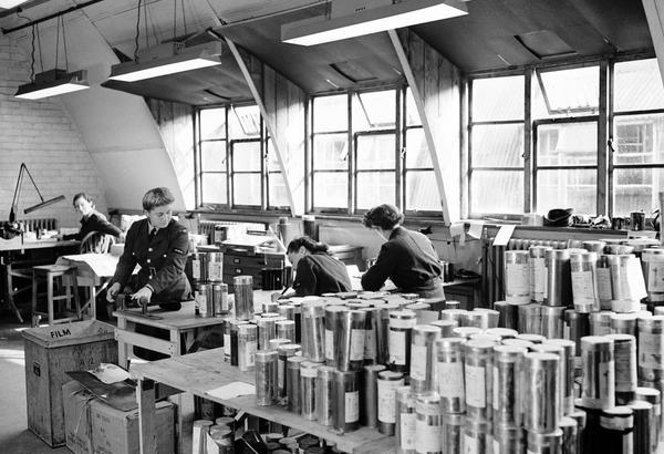 A black and white historic photo of 4 women in uniform working in an office. Large windows are visible in the background. 2 of the women face the camera, 2 are sitting with their backs to the camera. Metal film canisters cover almost every surface.