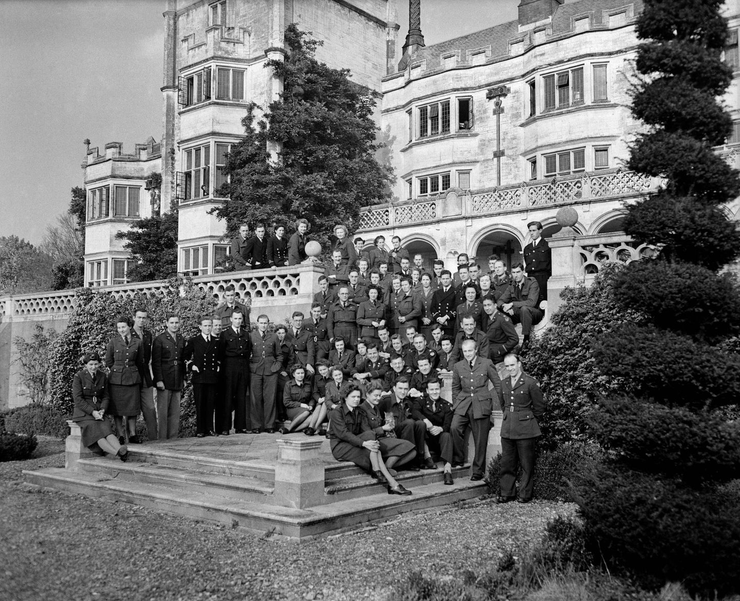 A black and white photo of a grand, white house. On the steps in front of the house is a big group of men and women wearing a variety of uniforms. There is a tree trimmed into a spiral shape in the right foreground.