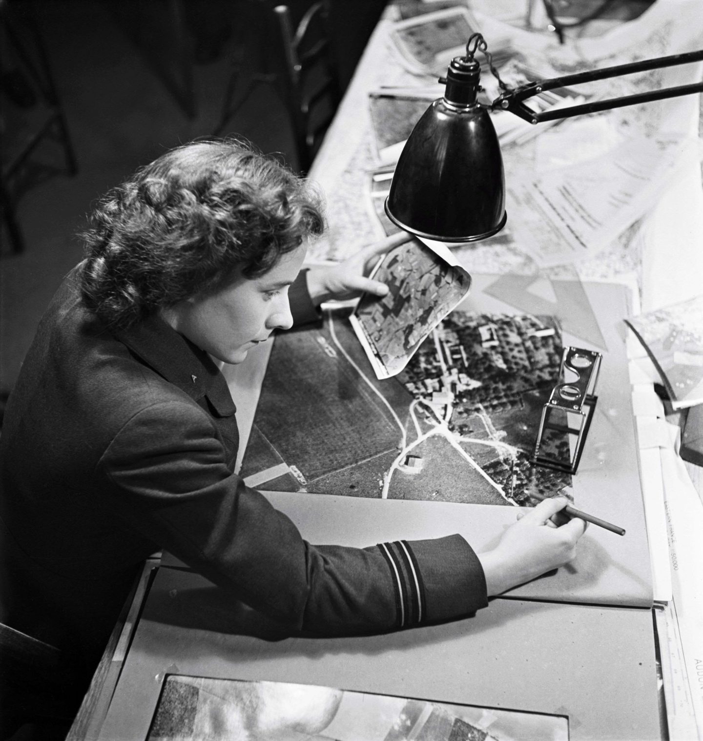 A black and white photo of a woman sitting at a desk. She is holding a small photo in her left hand and making notes on a larger photo with her right. A black lamp is suspended over the desk.