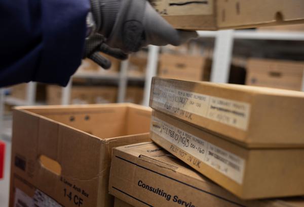 Two hands wearing grey gloves hold a slim cardboard box. They appear to be moving the box, and the bundle of boxes piled beneath it, on to the white shelving behind.