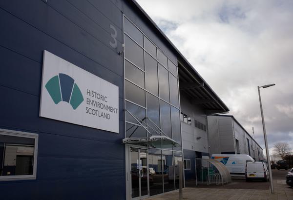 An industrial building with signage that shows a blue and teal logo. The sign reads ‘Historic Environment Scotland’. Above it is a grey ‘3’ – indicating the building unit number. The building is dark blue and has large glass windows. In the far right two white vans are parked.