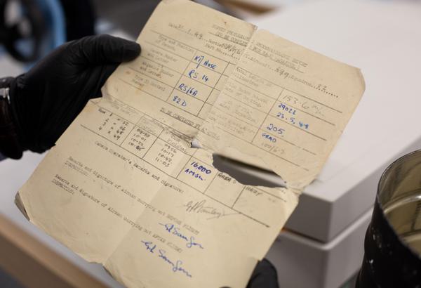 A close-up image of a piece of cream-coloured paper held by black gloved hands. It is torn on the centre fold lines and has creased and bent edges. It is printed with a form in black ink that has been completed in blue and grey handwritten text.
