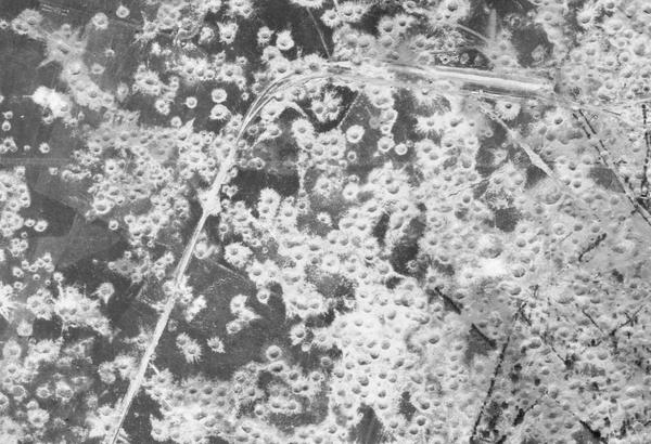 A black and white aerial photo of a very heavily bombed area of land. What land is visible is dark grey, and almost obscured with numerous white bomb craters. A road can be seen running from the bottom left corner to the top right.