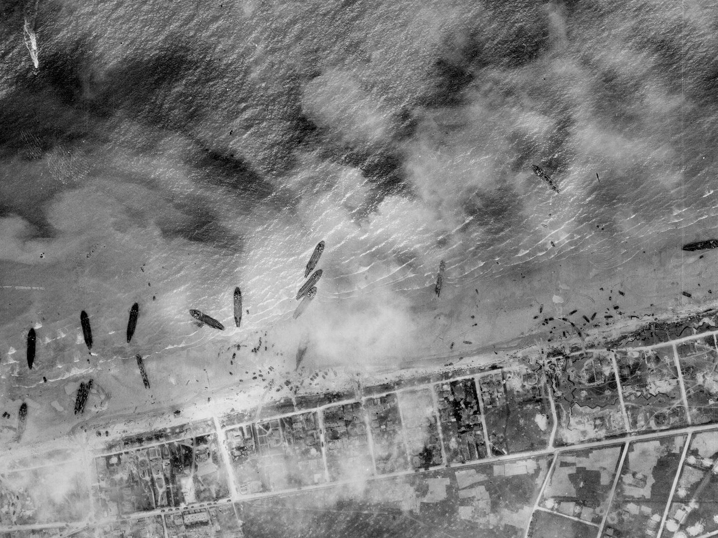 A black and white aerial photo of about a dozen ships pulled up on a beach. The land beyond the beach has been divided up into a grid, and the scene is slightly obscured by clouds or smoke.