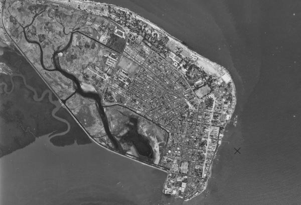 A black and white aerial photo of a city. The streets are arranged in a grid. There is a coastline to the right with an area of dark sea at the right-hand edge. To the left are two meandering watercourses and an area of undeveloped wetland.