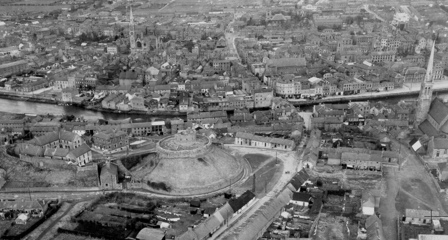 A historic black and white oblique aerial photo of Millmount Fort, Drogheda, a circular stone fort on a conical hill. The fort is located in a town, surrounded by houses and church spires, with a river running by.