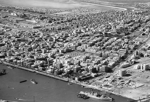 An oblique black and white aerial photo of a city with a coastline. The sea is at the bottom left corner. A small number of ships are on the water. The city is made up of what appears to be low-rise apartment blocks. There is a park in the centre.
