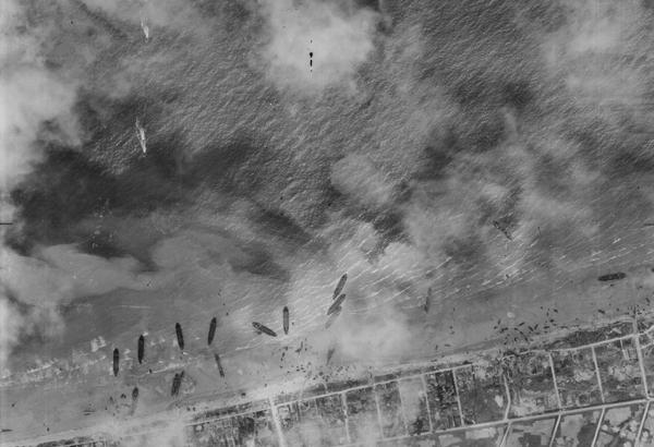 A black and white aerial photo of about a dozen ships pulled up on a beach. The land beyond the beach has been divided up into a grid, and the scene is slightly obscured by clouds or smoke.