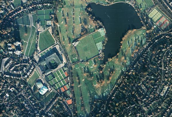 A colour aerial photo of the Wimbledon area in London. Streets of houses are visible at the right and left edges. A golf course with bunkers and trees occupies the middle. To the left of centre are about 20 tennis courts and a tennis stadium.