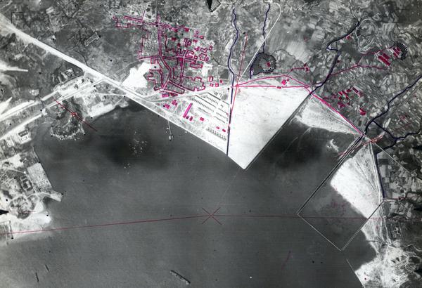 A black and white aerial photo of a city on the edge of the sea. There are buildings and an industrial area in the centre and fields to the right. The photo has been marked with purple pencil highlighting blocks of buildings.