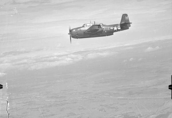A black and white oblique aerial photo of an airborne plane from side-on. The plane has one propellor and a see-through cockpit with the pilot visible. There is a rear-facing gun turret, and the fuselage is marked with an American star and ‘312’.