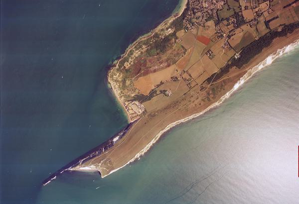 A colour aerial photo of the sea, very dark blue to the left and white from the glare of the sun on the right. In the middle a thin peninsula covered in brown grass extends into the sea. It has white cliffs and ends in a series of individual stacks.
