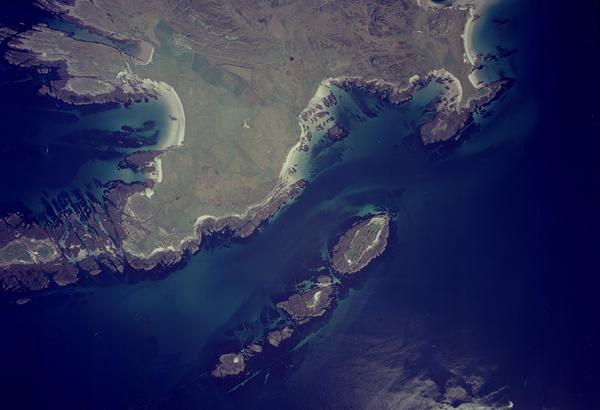 A colour aerial photo of islands surrounded by a deep blue sea. There is one big island left of centre and a small one to the right. The islands have green grass and a mix of rocky and sandy shores. The sea is light blue over the sand.