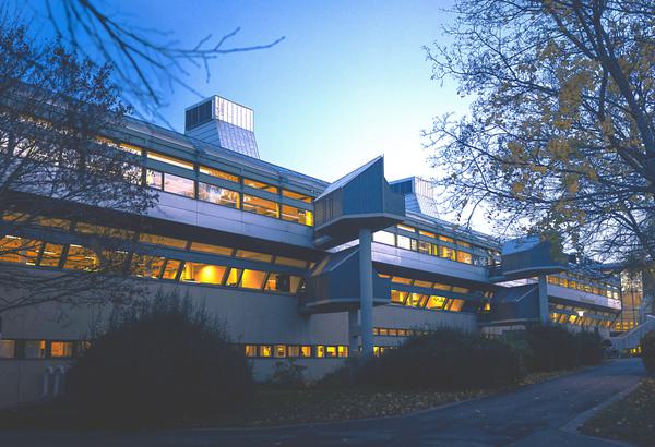 A long, low profile building fills the frame. It has lots of square edges and sharp angles, and our view of it is partially blocked by spindly trees and dense, low bushes. The photograph has been taken at night, and the clear, dark blue sky glows behind the building. Three rows of windows are illuminated with orange light from inside.