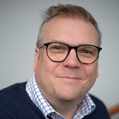 A head-and-shoulders view of a smiling man with white skin and swept-back grey hair, wearing dark-framed glasses, a blue checked shirt and a dark blue jumper.