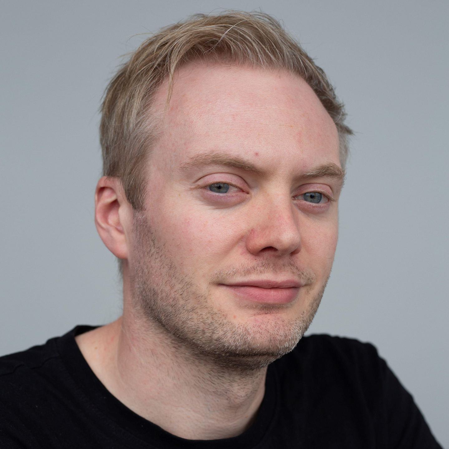 A head-and-shoulders view of a man with white skin, stubble and swept-back blonde hair, wearing a black top.