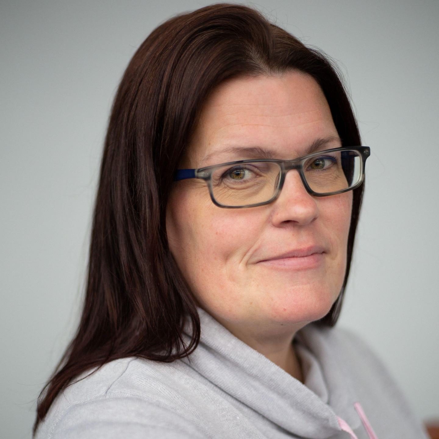 A head-and-shoulders view of a smiling woman with white skin and long dark hair hanging loose, wearing grey-framed glasses and a light grey jumper.