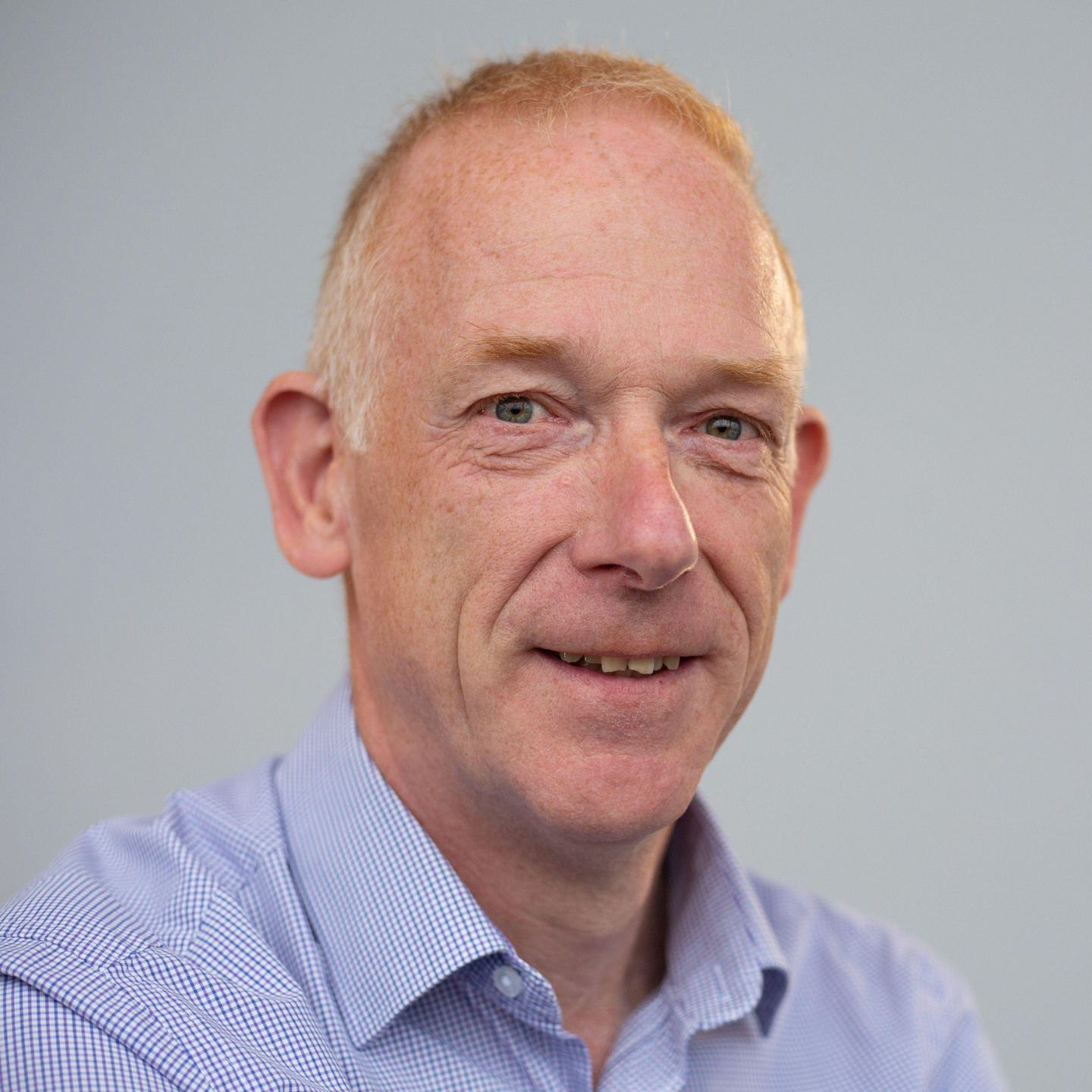 A head-and-shoulders view of a smiling man with white skin and short red hair, wearing a light blue shirt.